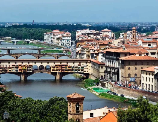 A panoramic view of Florence featuring the Ponte Vecchio bridge over the Arno River, surrounded by historic buildings and greenery.