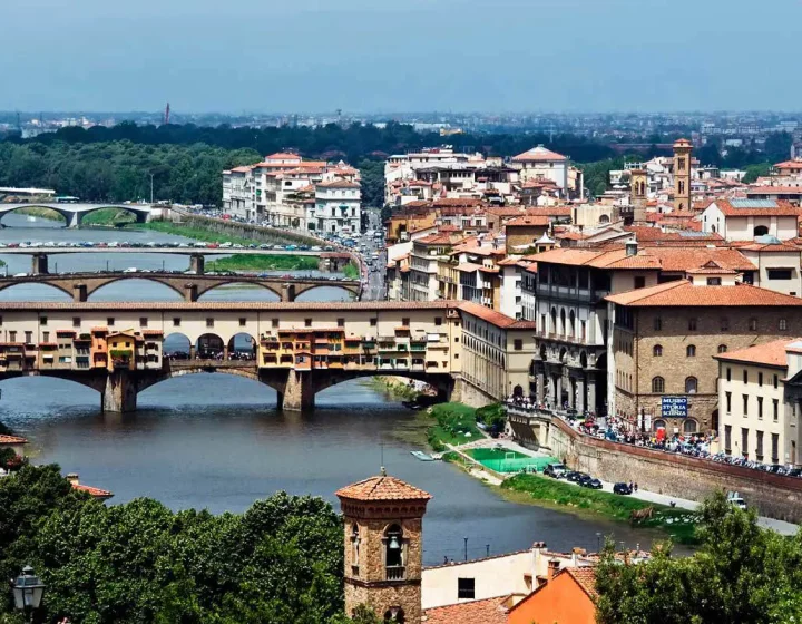 A panoramic view of Florence featuring the Ponte Vecchio bridge over the Arno River, surrounded by historic buildings and greenery.